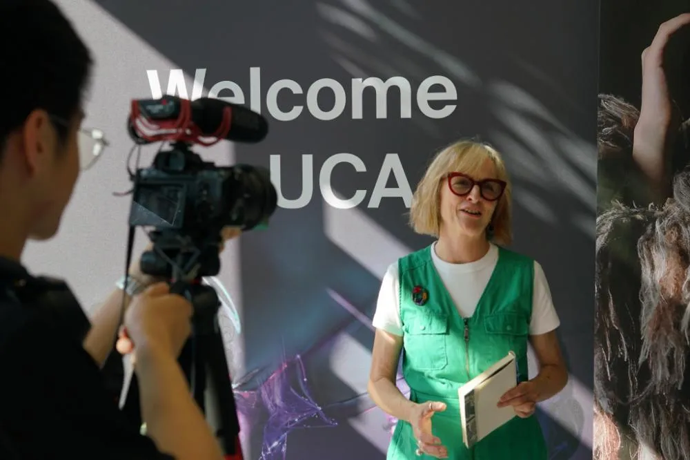 A camera operator films a presenter in a green vest holding a book in front of a Welcome UCA backdrop SUSAN ORR