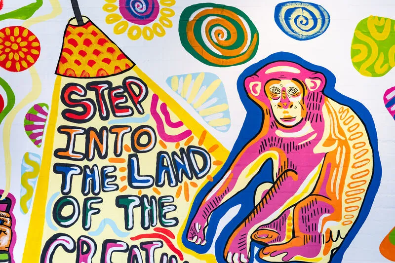 An illustration of a blue, yellow and pink monkey on a white wall, next to a red and yellow lamp shining a light on 'Step into the land of the' written underneath. The next word in the sentence is cut off from the photo.