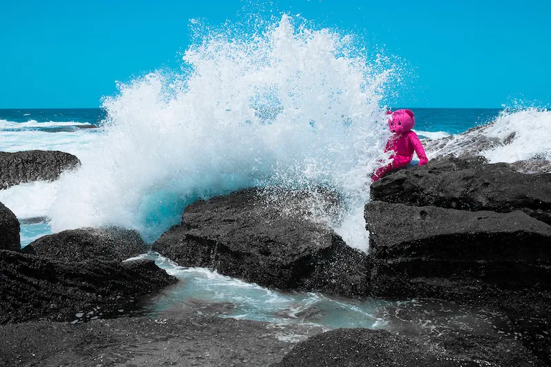 A photo by LUAP showing a giant pink bear sitting on a rock with a wave crashing over it, entitled 