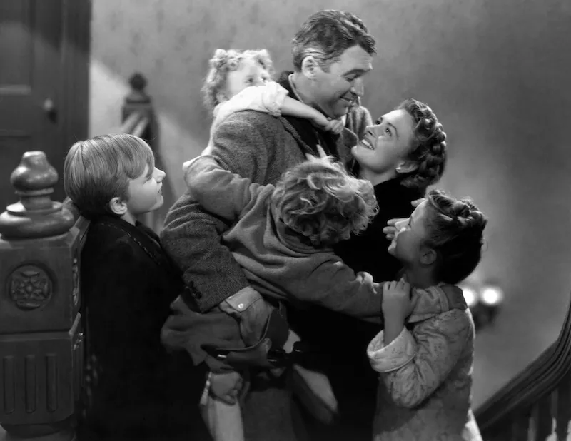 Image is a still from the Christmas film, It's a Wonderful Life, showing the main character surrounded by his family.