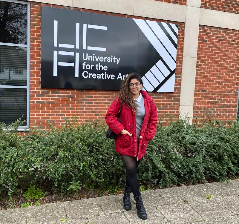 International student Sasha Miranda, in a red coat, stands in front of some UCA signage at the Epsom campus