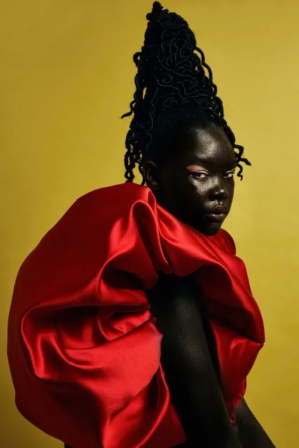 Image shows a black woman dressed in red satin against a yellow background, her hair styled above her head