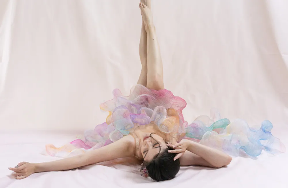 A model wears a pastel chiffon textile dress. She is lying on the floor, arms stretched out beside her and her legs in the air, replicating a balletic pose