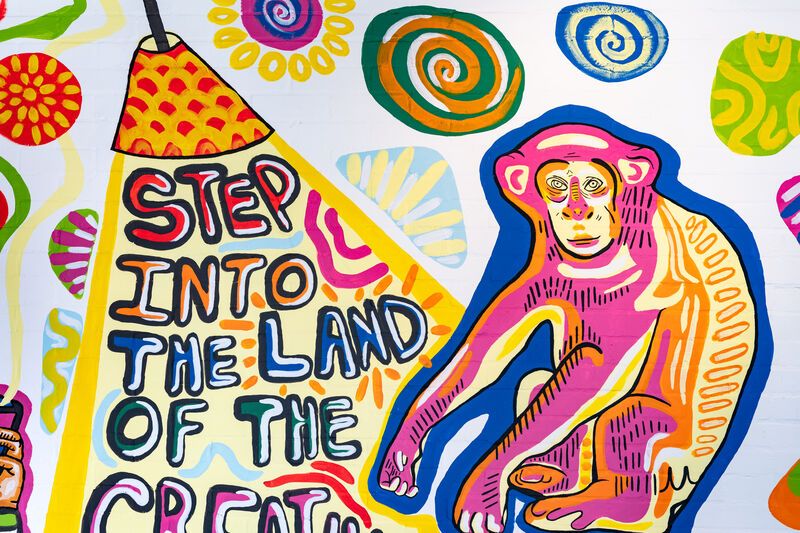 An illustration of a blue, yellow and pink monkey on a white wall, next to a red and yellow lamp shining a light on 'Step into the land of the' written underneath. The next word in the sentence is cut off from the photo.